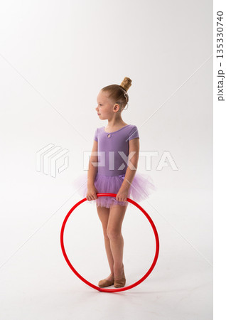 A smiling ballerina with a cochlear implant raises a red hoop in a ballet pose. Inclusive sport, hearing technology, and confident childhood expression through rhythmic dance and movement. 135330740