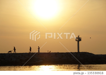 Dawn at sea.A golden sunrise over the sea. A stone pier with tourists and a lighthouse. 135330918