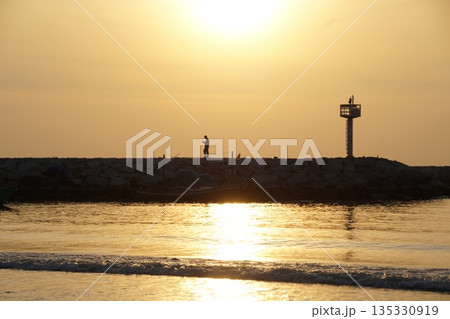 A golden sunrise over the sea. A stone pier with tourists and a lighthouse.Dawn at sea. A golden sunrise over the sea. A stone pier with tourists and a lighthouse.Dawn at sea. 135330919