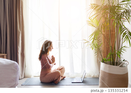 Adult woman practicing seated yoga meditation with laptop nearby in hotel room Adult woman practicing seated yoga meditation with laptop nearby in hotel room 135331254