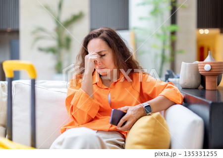Stressed woman holding smartphone and touching face while sitting in hotel lounge Stressed woman holding smartphone and touching face while sitting in hotel lounge 135331255