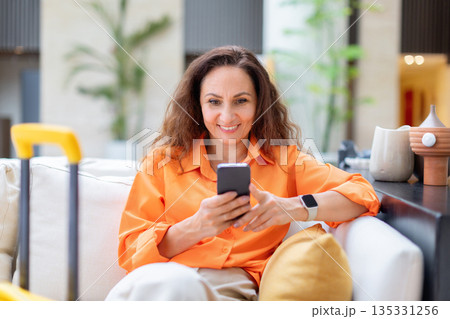 Smiling adult woman sitting in hotel lobby using smartphone near yellow suitcase 135331256