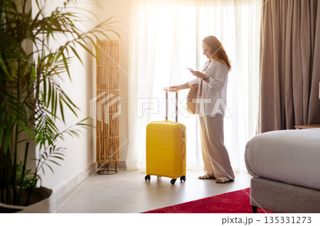 Adult woman checking smartphone while standing with yellow suitcase in hotel room 135331273