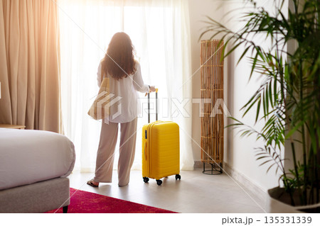 Adult woman standing near hotel window with suitcase and bag, looking outside 135331339