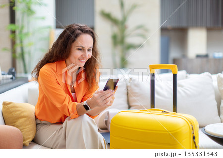 Smiling adult caucasian woman using smartphone beside luggage in hotel lounge 135331343