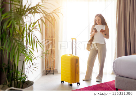 Adult caucasian woman checking smartphone and passport near suitcase in hotel room 135331396