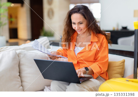 Adult woman working with laptop and documents while sitting in hotel lobby Adult woman working with laptop and documents while sitting in hotel lobby 135331398