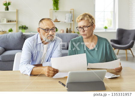 Senior couple reviewing bills and finances at home with tablet and papers Senior couple reviewing bills and finances at home with tablet and papers 135331957