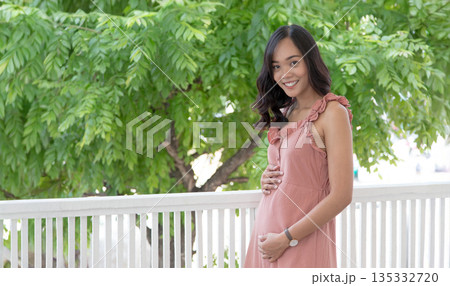 Happy pregnant Asian woman in pink dress touching her belly in the garden. Concept of healthy pregnancy, maternity care, and expectation of a new life with nature Happy pregnant Asian woman in pink dress touching her belly in the garden. Concept of healthy pregnancy, maternity care, and expectation of a new life with nature 135332720