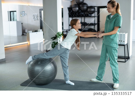 Pediatric physiotherapist helping child balance on therapy ball 135333517