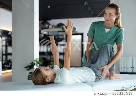 Young patient practicing movement using Pilates ring under supervision Young patient practicing movement using Pilates ring under supervision 135333519