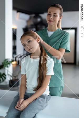 Woman therapist performing soft neck stretching little girl sitting on couch 135334098