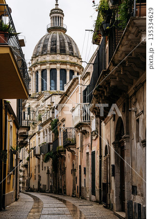 Back Side Of Duomo Di San Giorgio In Ragusa, Sicily: Narrow Historic Street Leading Toward Baroque Cathedral Dome And Stone Facades 135334629