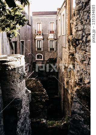 Teatro Romano E Odeon In Catania, Sicily: Ancient Roman Theater Ruins With Stone Seating, Historic Buildings, And Church Towers Under Clear Daylight 135334685