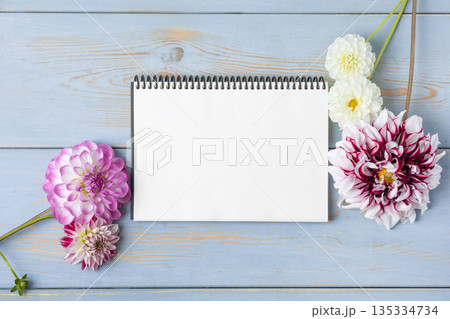 Notebook with clean page with purple flowers dahlias on blue wooden background. Colorful flower background with space for text. Nature trendy decorative design. Flat lay. Notebook with clean page with purple flowers dahlias on blue wooden background. Colorful flower background with space for text. Nature trendy decorative design. Flat lay. 135334734