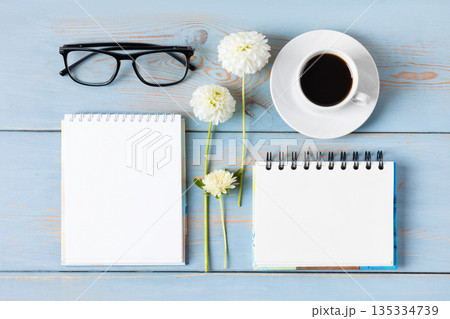 Top view of the desktop. Spiral notebook with an open blank page, glasses, pen, white cup with black coffee, dahlia flowers. Festive office desktop concept. Morning coffee cup. Top view of the desktop. Spiral notebook with an open blank page, glasses, pen, white cup with black coffee, dahlia flowers. Festive office desktop concept. Morning coffee cup. 135334739