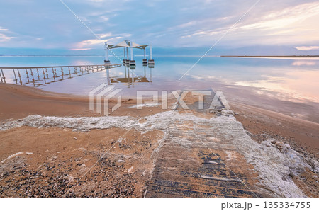 Calm morning at Ein Bokek Dead Sea beach, pink clouds reflection over water surface near steel rails leading to sun shade shelter, sand covered with salt crystals 135334755