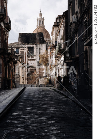 Narrow Street In Catania With Church Dome: Italian Sicilian City Scene With Historic Stone Buildings, Strong Light Contrast, And Deep Urban Perspective Narrow Street In Catania With Church Dome: Italian Sicilian City Scene With Historic Stone Buildings, Strong Light Contrast, And Deep Urban Perspective 135334782