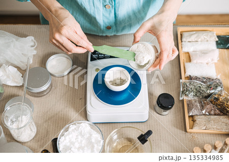 Hands scoop thick white base into small bowl on digital scale surrounded by laboratory equipment and jars. Consumer centric products, DIY beauty trends, niche market manufacturing. 135334985
