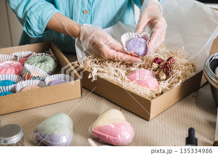 Person wearing protective gloves packs handmade purple bath bomb into gift box filled with shredded paper. Artisanal business, home-based startup, craft industry 135334986
