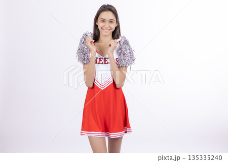 Cheerful cheerleader girl holding pompons and smiling for team celebration Cheerful cheerleader girl holding pompons and smiling for team celebration 135335240