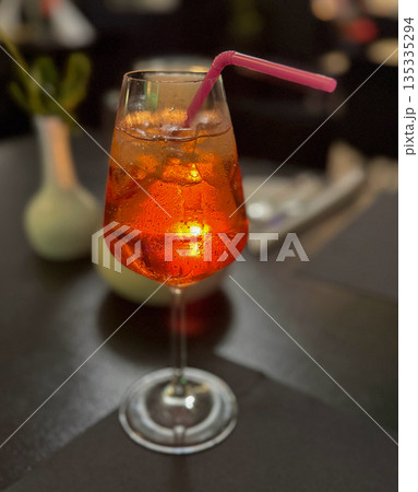 Glowing orange cocktail with ice in a wine glass on a dark table. Warm evening light creates a cozy bar mood Glowing orange cocktail with ice in a wine glass on a dark table. Warm evening light creates a cozy bar mood 135335294