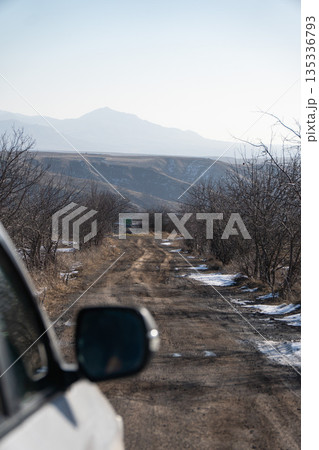 Rural dirt road in Armenia seen from car window, with bare trees, patches of snow, distant mountains creating a quiet road trip mood Rural dirt road in Armenia seen from car window, with bare trees, patches of snow, distant mountains creating a quiet road trip mood 135336793