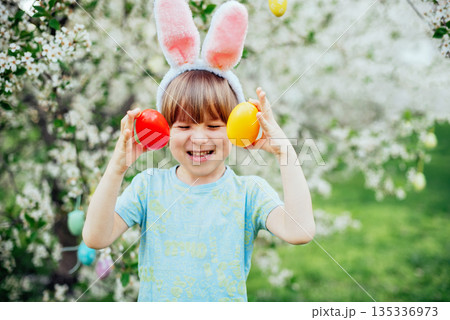 Easter Egg Hunt with Child Carrying Bright Plastic Eggs. Smiling child wearing bunny ears in a blooming spring garden Easter Egg Hunt with Child Carrying Bright Plastic Eggs. Smiling child wearing bunny ears in a blooming spring garden 135336973