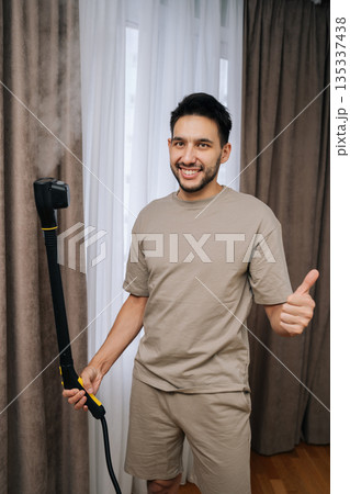 Vertical portrait of cheerful man showing thumbs up, providing professional curtain steam cleaning service for hygienic home environment, ensuring freshness and cleanliness with specialized equipment. 135337438