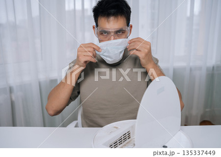 Front view of young man wearing safety glasses and holding face mask, preparing to clean robot vacuum cleaner, ensuring personal protection during home appliance maintenance. 135337449