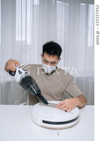 Vertical portrait of technician male wearing protective face mask and goggles cleaning dirty filter from robot vacuum cleaner using small handheld vacuum for optimal device maintenance. 135337452
