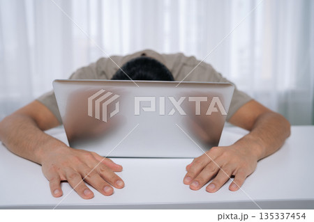 Exhausted man feeling burnout and stress, laying sleeping with face on laptop after working long hours, experiencing fatigue and overwork in workplace or home office. 135337454