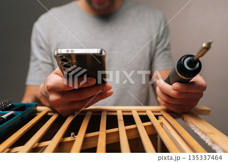 Cropped shot of handyman assembling wooden furniture, holding cordless electric screwdriver and checking instructions on smartphone for diy home improvement and renovation project. Cropped shot of handyman assembling wooden furniture, holding cordless electric screwdriver and checking instructions on smartphone for diy home improvement and renovation project. 135337464