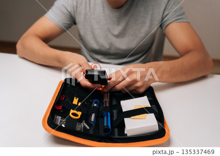 Person handling multimeter and organizing various small tools inside compact orange and black zippered case, preparing for or performing electronics repair and assembly tasks. Person handling multimeter and organizing various small tools inside compact orange and black zippered case, preparing for or performing electronics repair and assembly tasks. 135337469