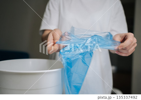 Hands unfolding and positioning fresh blue plastic bag into empty white trash can, signifying start of efficient waste management and cleanliness routine in domestic setting, close-up cropped shot. Hands unfolding and positioning fresh blue plastic bag into empty white trash can, signifying start of efficient waste management and cleanliness routine in domestic setting, close-up cropped shot. 135337492
