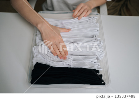 Close-up top view of hands organizing freshly folded white and black socks in transparent storage container, demonstrating efficient organization and tidiness for home wardrobe management. Close-up top view of hands organizing freshly folded white and black socks in transparent storage container, demonstrating efficient organization and tidiness for home wardrobe management. 135337499