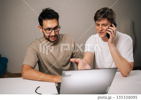 Portrait of two two young startup men brainstorming startup ideas, collaborating ona new business project, using laptop and smartphone for communication and strategy in modern home office. 135338095