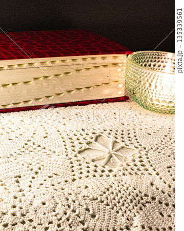 Red book with gilt page edges on a crocheted white tablecloth. Contemplative scene Red book with gilt page edges on a crocheted white tablecloth. Contemplative scene 135339561