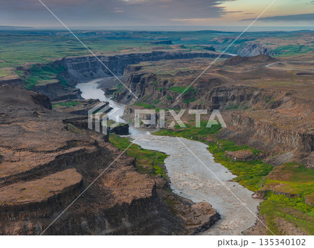 Studlagil Canyon in Iceland features dramatic basalt columns, a winding glacial river, earthy canyon walls, green vegetation, and a cloudy sky. Studlagil Canyon in Iceland features dramatic basalt columns, a winding glacial river, earthy canyon walls, green vegetation, and a cloudy sky. 135340102