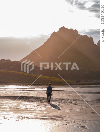 A person walks on wet sand reflecting golden light, with jagged mountains silhouetted in the background near Iceland's Stokknes Peninsula. A person walks on wet sand reflecting golden light, with jagged mountains silhouetted in the background near Iceland's Stokknes Peninsula. 135340110