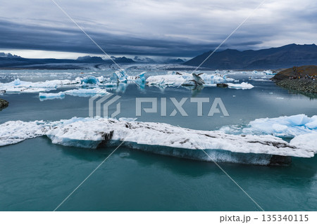 Large icebergs float in turquoise water at Jokulsarlon Glacier Lagoon, framed by mountains and an overcast sky. People stand on the rocky shoreline. Large icebergs float in turquoise water at Jokulsarlon Glacier Lagoon, framed by mountains and an overcast sky. People stand on the rocky shoreline. 135340115