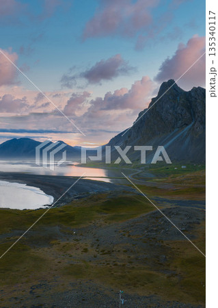 The Stokksnes Peninsula in Iceland features a mountain range, black sand beach, and reflective water under soft sunset hues, creating a serene scene. 135340117