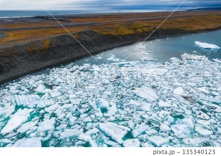 Aerial view of a glacial lagoon in Iceland featuring floating icebergs, turquoise water, a rugged shoreline, a road, and the open sea on the horizon. Aerial view of a glacial lagoon in Iceland featuring floating icebergs, turquoise water, a rugged shoreline, a road, and the open sea on the horizon. 135340123