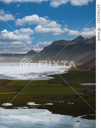 Aerial view of Stokknes Peninsula in Iceland, showing rugged peaks, serene coastline, reflective water, green fields, and a partly cloudy sky. 135340125