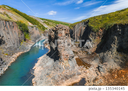 Studlagil Canyon in Iceland features dramatic basalt columns, a vibrant turquoise river, lush green hills, and a clear blue sky in a volcanic landscape. 135340145