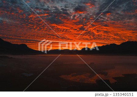 A vibrant red and orange sunset illuminates the sky over silhouetted Icelandic peaks. Mist blankets the landscape, with reflections in a tranquil water surface. A vibrant red and orange sunset illuminates the sky over silhouetted Icelandic peaks. Mist blankets the landscape, with reflections in a tranquil water surface. 135340151
