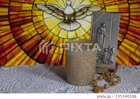 Religius items in the foreground with blurred stained glass dove of the Holy Spirit evoking faith, devotion and hope Religius items in the foreground with blurred stained glass dove of the Holy Spirit evoking faith, devotion and hope 135340186