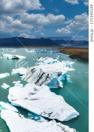 Aerial view of Jokulsarlon Glacier Lagoon in Iceland, featuring white icebergs in turquoise water, rugged mountains, and a clear blue sky with clouds. 135340189