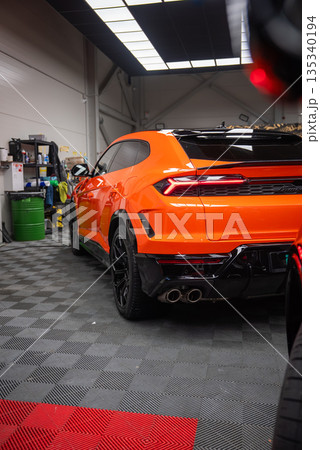 A three quarter rear view of an orange Lamborghini Urus SE parked indoors, showing sharp taillights, quad exhaust tips, black diffuser, and gloss black wheels. A three quarter rear view of an orange Lamborghini Urus SE parked indoors, showing sharp taillights, quad exhaust tips, black diffuser, and gloss black wheels. 135340194