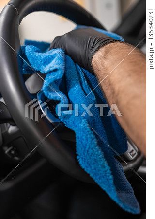 A black nitrile gloved hand wipes a car steering wheel with a bright blue microfiber towel inside a modern vehicle, dashboard controls visible, shallow depth of field. A black nitrile gloved hand wipes a car steering wheel with a bright blue microfiber towel inside a modern vehicle, dashboard controls visible, shallow depth of field. 135340231